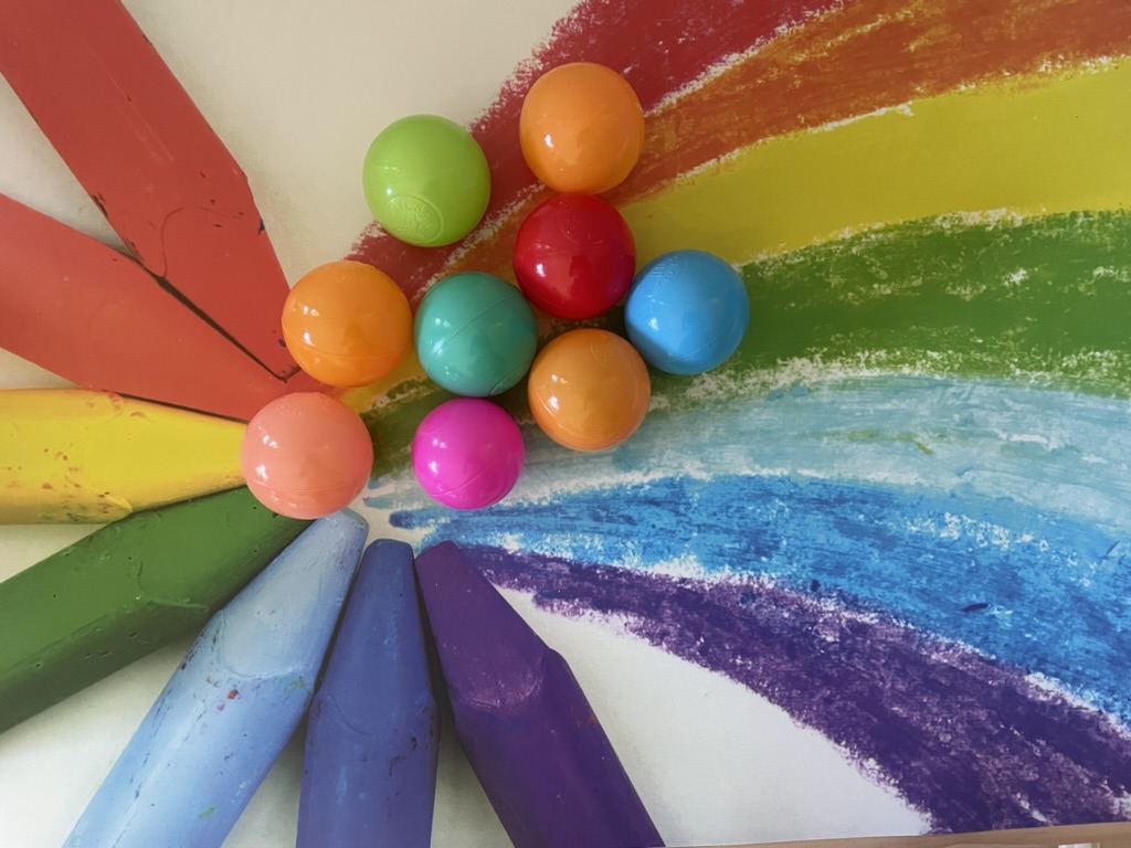 Un ensemble de balles colorées et de gros crayons pastels disposés sur un dessin d'arc-en-ciel, créant une ambiance ludique et créative.